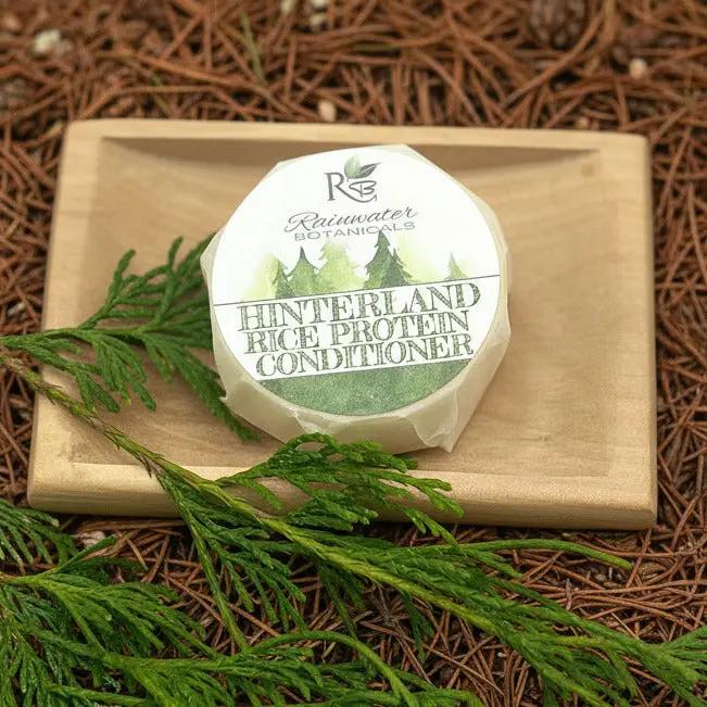 Round container of Rainwater Botanicals Hinterland Rice Protein Conditioner on a wooden block with greenery.