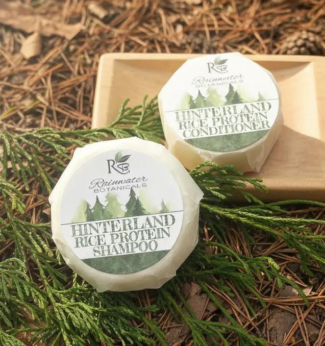 Two containers of Hinterland Rice Protein Shampoo and Conditioner from Rainwater Botanicals on a white background with greenery.