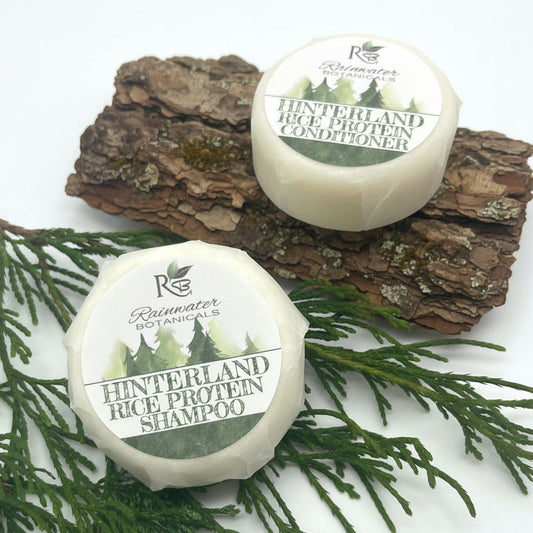 Two containers of Hinterland Rice Protein Shampoo and Conditioner from Rainwater Botanicals on a wooden tray with greenery.
