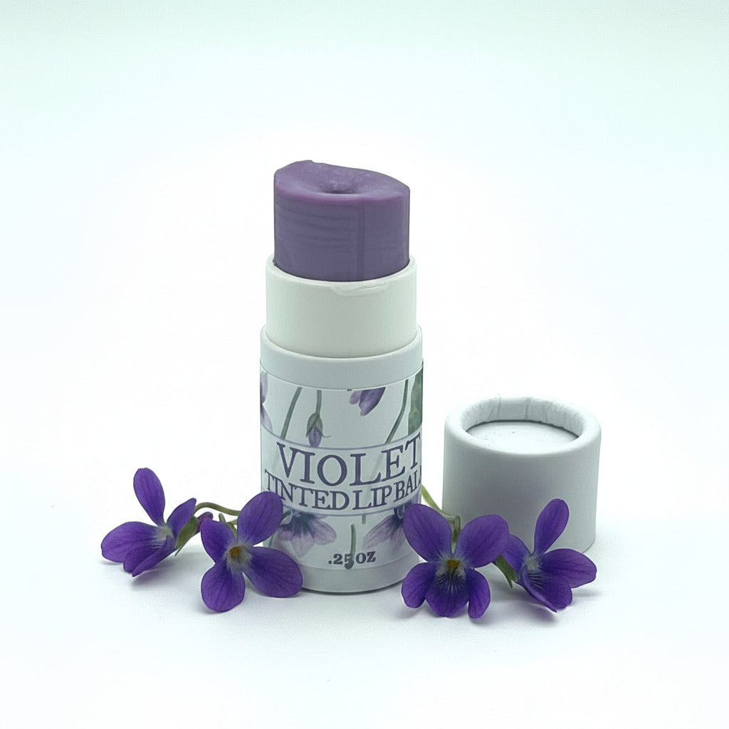 A tube of violet tinted lip balm with its cap removed and placed beside it on a clean white background. The tube features a white label with purple flowers and the text 'VIOLET TINTED LIP BALM .