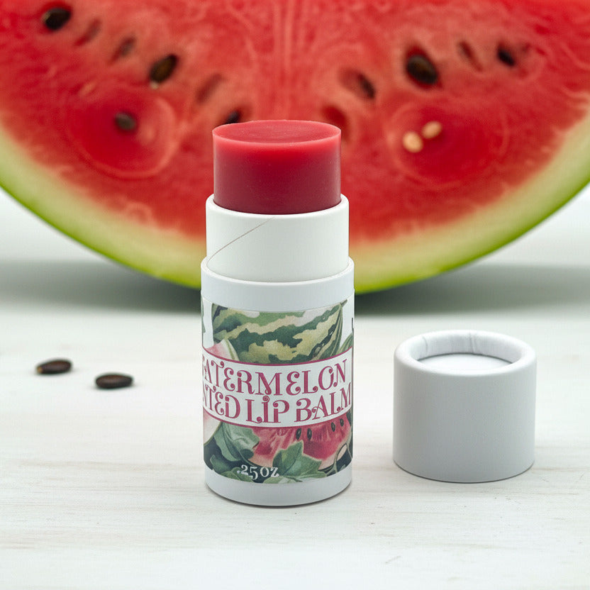 A tube of watermelon flavored lip balm with its cap removed and placed beside it on a clean white background. The balm stick has a red top, and the label features watermelon illustrations with the text 'WATERMELON FLAVORED LIP BALM'.