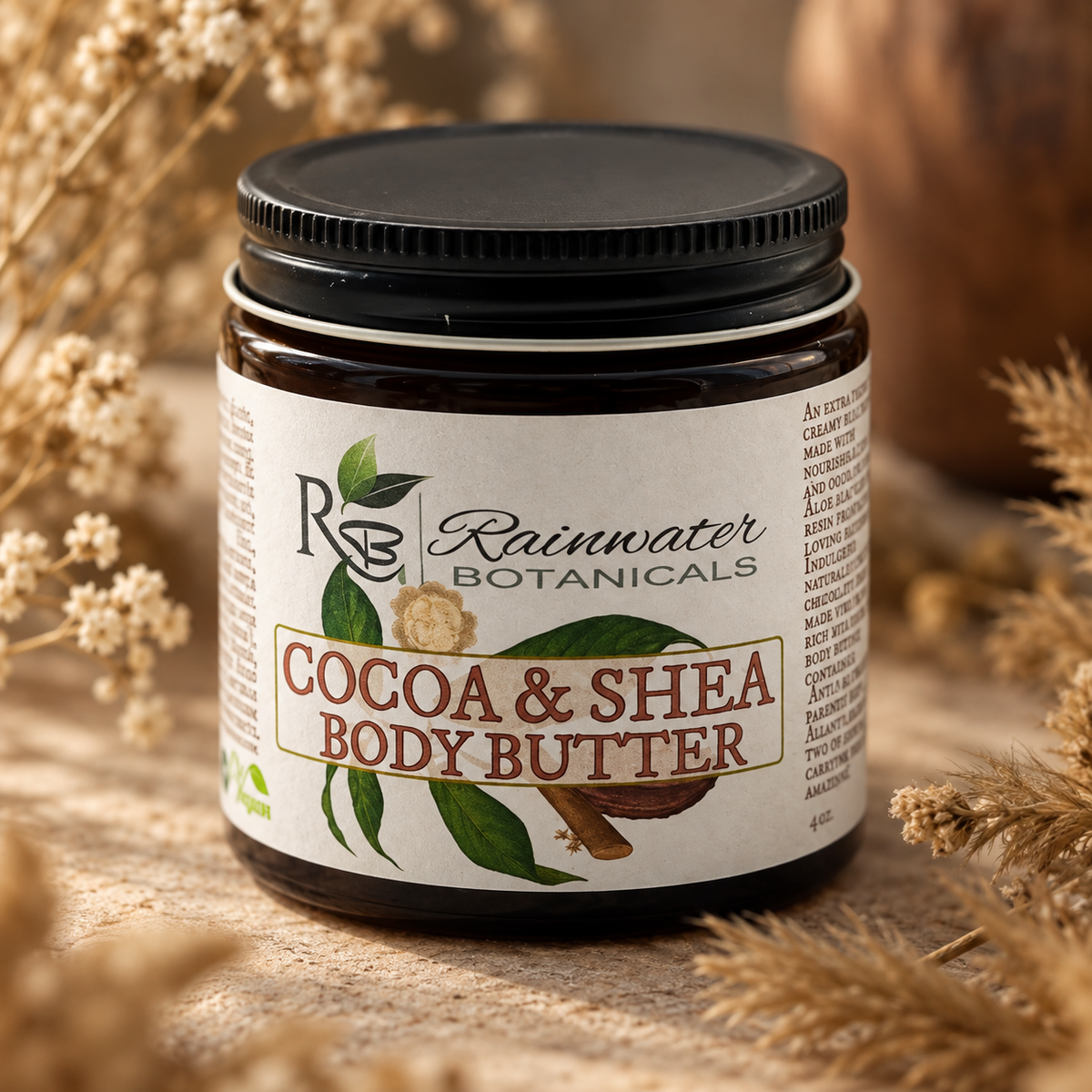 A jar of Rainwater Botanicals Cocoa & Shea Body Butter, featuring a black lid and a cream-colored label with green leaf illustrations, set against a rustic background of dried wheat and wood.