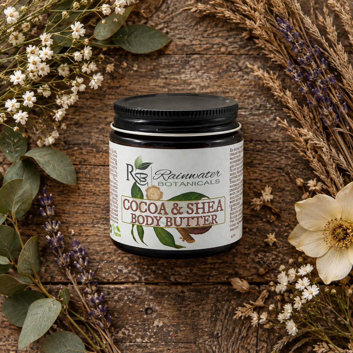 A jar of Rainwater Botanicals Cocoa & Shea Body Butter sits on a rustic wooden surface, surrounded by dried flowers, green leaves, and lavender sprigs.