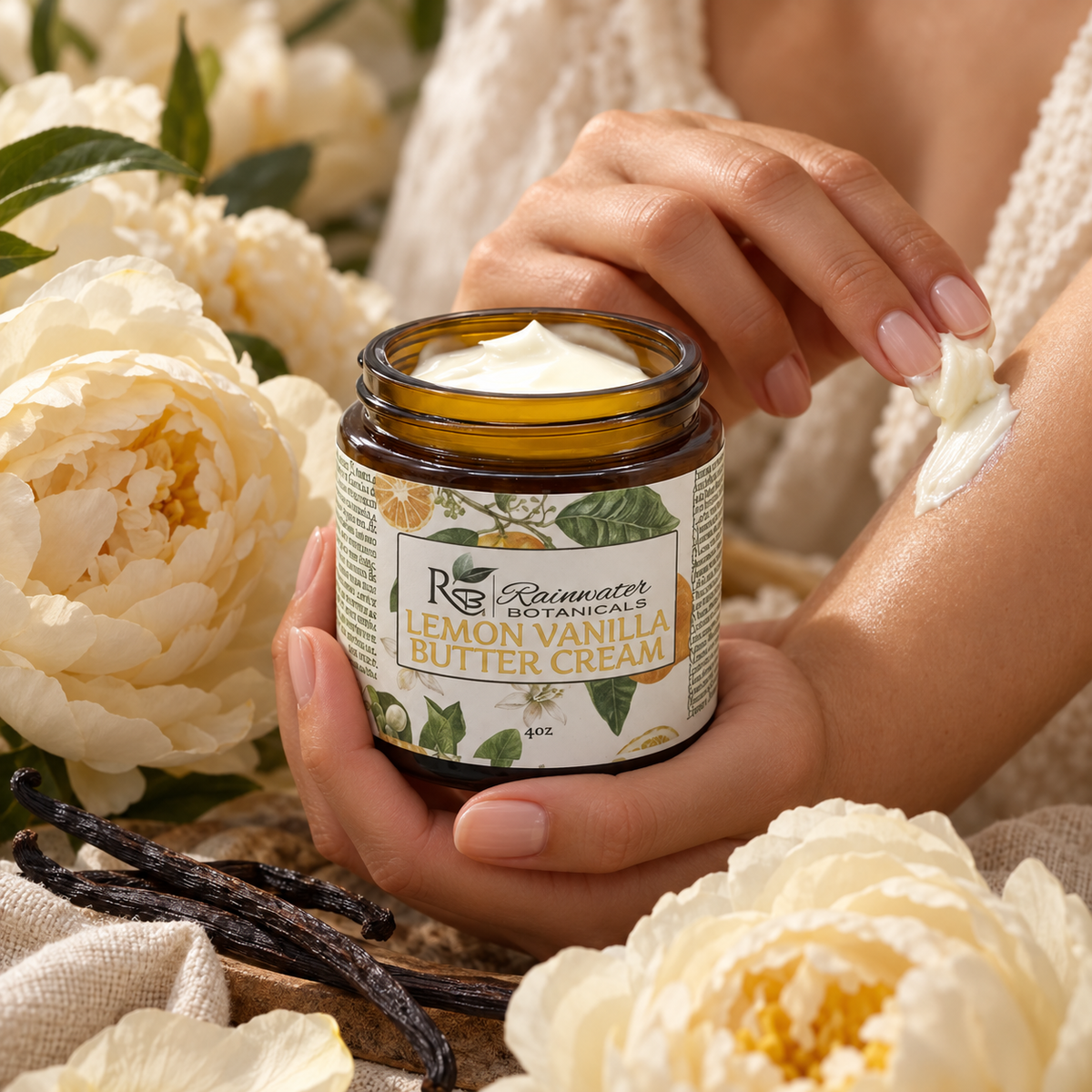 A person holds an amber glass jar of Rosewater Naturals Lemon Vanilla Butter Cream, with white cream being spread onto their arm. The scene is decorated with white peony flowers and vanilla beans.